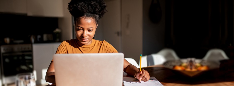 Mulher faz anotações em caderno enquanto estuda pelo computador em sua casa