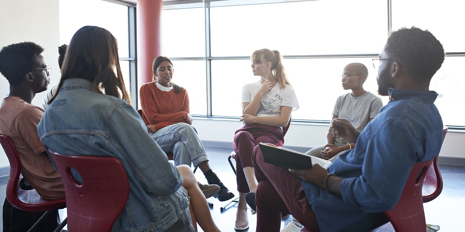 Depressed young woman sharing while sitting with friends and mental health instructor in group meeting at classroom